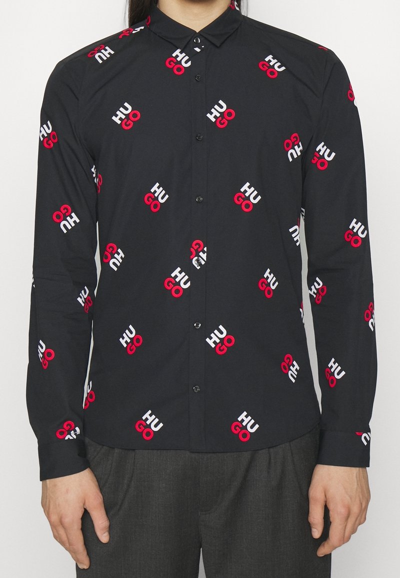 Czarna koszula na guziki z powtarzającym się nadrukiem logo "HUGO" w kolorach czerwonym i białym, wykonana z gładkiej tkaniny, z klasycznym kołnierzykiem i długimi rękawami.