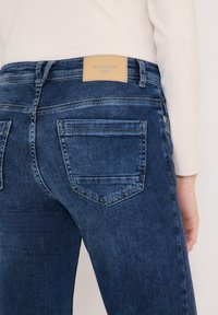 Tmavě modré džínové kalhoty s hladkou texturou, viditelným prošitím a koženou záplatou na pase. Mají dvě zadní kapsy.