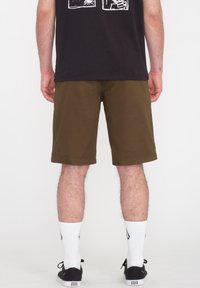 Braune Shorts mit glatter Textur, die bis zur Mitte des Oberschenkels reichen. Getragen mit weißen Socken und schwarzen Schuhen, die einfache Designelemente aufweisen.
