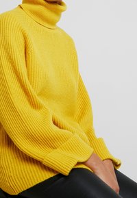 Žlutý žebrovaný rolák s dlouhými rukávy a volným střihem, s texturovaným pleteným vzorem, zkombinovaný s černými koženými kalhotami.