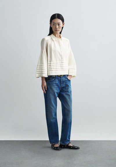 Kvinde stående med hænder i lommerne iført cremet knap-op-bluse med plisserede klokkeærmer, blå jeans og sorte flade sko med nitter på gråt gulv.