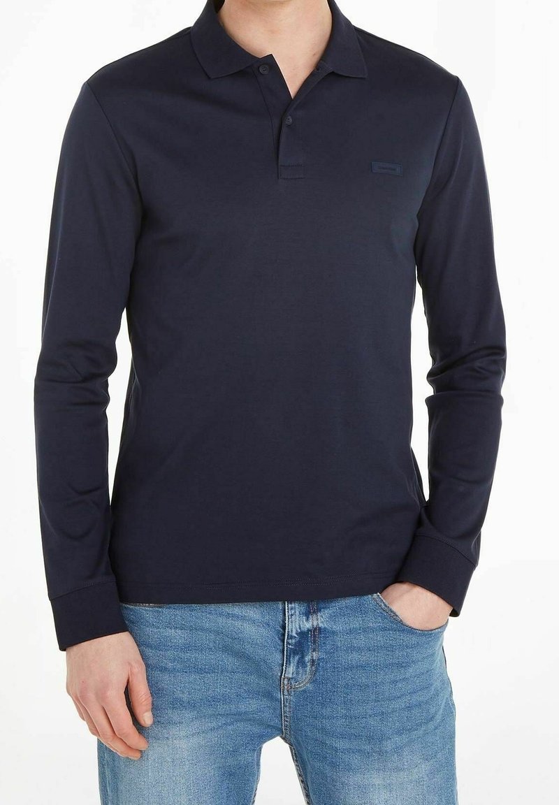 Marinblå långärmad polo-shirt i mjuk bomull, med knäppt krage, slim fit och diskret logotyp på bröstet. Bärs med denimjeans.