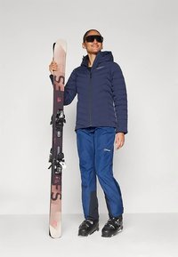 Inimene, kes kannab suusavarustust, hoides püstiselt suuski, riietatud tumesinisesse jope, sinistesse pükstesse, suuskade saapaisse ja suurtesse tumedatesse prillidesse, naeratab.