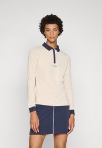 Long-sleeve, light beige polo shirt with a navy collar and cuffs. Features a small embroidered logo on the chest. Worn with a navy skirt.