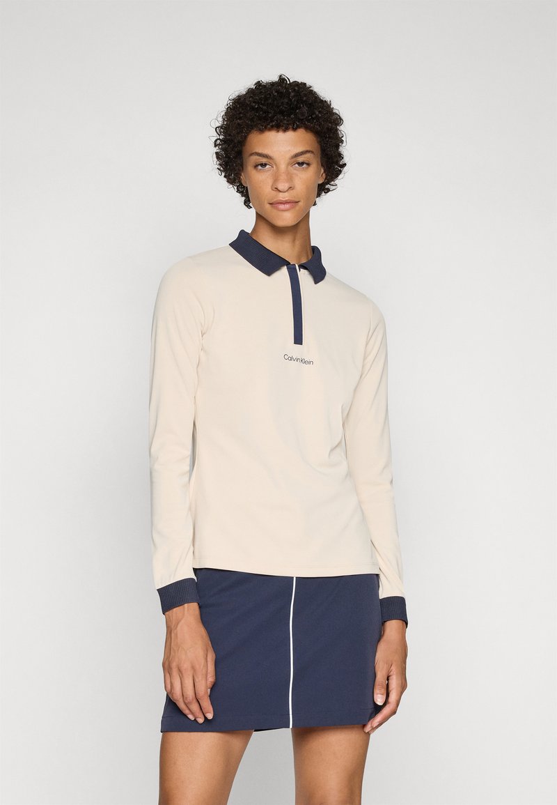 Long-sleeve, light beige polo shirt with a navy collar and cuffs. Features a small embroidered logo on the chest. Worn with a navy skirt.