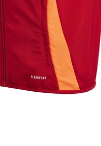 Rote und orange Sportjacke mit glatter Textur, ausgestattet mit einem seitlichen Paneeldesign und "AEROREADY"-Logo nahe dem Saum. Mit Reißverschluss vorne.