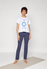 Bílé krátké tričko s modrým motivem slunce a nápisem, v kombinaci s přiléhavými tmavě modrými legínami. Modelka stojí bosá na světle zbarvené podložce.