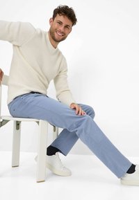 Maglione chiaro e texturizzato con collo a giro; pantaloni slim-fit azzurri; sneakers bianche; seduto su una sedia bianca contro uno sfondo neutro.