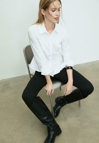 Weißes, tailliertes Hemd, kombiniert mit schwarzen Skinny-Jeans und kniehohen schwarzen Lederstiefeln. Silberne Ohrringe und ein Armband sind ebenfalls zu sehen.