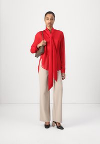 Rød gjennomsiktig bluse med drapert halslinje, beige vide bukser og svarte spisse hæler. Modellen holder en liten brun veske.