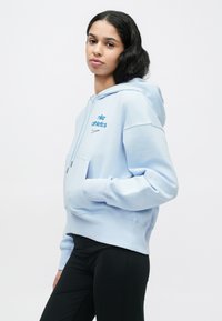 Φούτερ φούξια ανοιχτού μπλε χρώματος με μπροστινή τσέπη και κεντημένο λογότυπο "nike athletics", με ριμπ μανσέτες και τελείωμα. Φορεμένο με μαύρα κολάν.