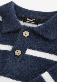 Polo di misto lana blu navy con finitura testurizzata, strisce orizzontali bianche e bottoni beige. L'etichetta riporta "NEXT, Made in Myanmar."