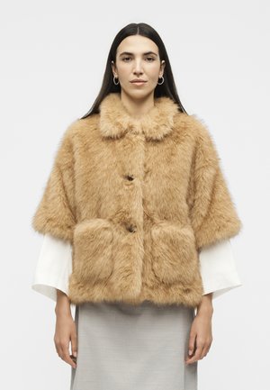 Femme aux longs cheveux foncés, portant des boucles d'oreilles créoles en or et une veste courte en fausse fourrure beige sur un haut blanc et une jupe gris clair.