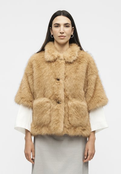 Femme aux longs cheveux foncés, portant des boucles d'oreilles créoles en or et une veste courte en fausse fourrure beige sur un haut blanc et une jupe gris clair.