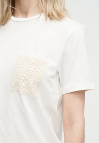 Κοντινό πλάνο ενός λευκού πουκαμίσου με κοντό μανίκι και υφασμένο, διακοσμητικό τσέπη από εκρού δαντέλα στο στήθος.