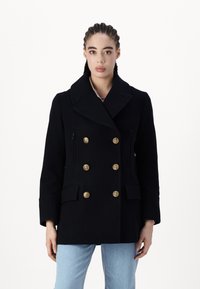 Mujer con trenzas que lleva un abrigo negro de doble botonadura con botones dorados y jeans azul claros, de pie frente a un fondo blanco.