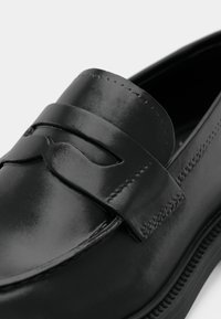Mocassin en cuir noir avec une finition lisse, des détails cousus et une sangle décorative sur le devant. Bout arrondi et design minimaliste.