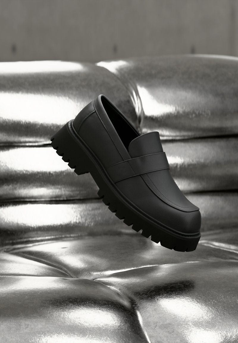 Μαύρο μοκασίνι από δέρμα με χοντρή σόλα τύπου τρακτέρ που αιωρείται πάνω από μια γυαλιστερή ασημένια επιφάνεια με καπιτονέ, αναδεικνύοντας το σχήμα και την υφή του.