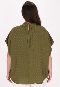 Oliwkowa bluzka o luźnym kroju, z krótkimi rękawami i detalem w postaci wiązania z tyłu. Posiada teksturowany wzór z małymi, wypukłymi kropkami.