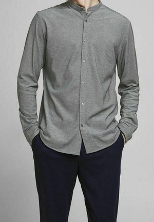 Mann trägt ein graues, geknöpftes Hemd mit Mandarin-Kragen und langen Ärmeln sowie dunkle Hose, die Hände in den Taschen, steht vor einem einfarbigen Hintergrund.