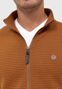 Homme portant une veste zippée marron texturée par-dessus un t-shirt blanc à col en V, avec une fermeture éclair visible et un badge rond avec logo sur la poitrine.