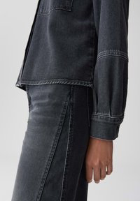 Seitenansicht einer Person in einer schwarzen Jeansjacke und passenden hochgeschnittenen Jeans, mit sichtbaren Nähten und entspanntem Händedruck.