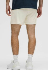 Cremefarvede shorts lavet af blødt stof, der har et tætsiddende design, sidesømme og et lille logo nær kanten. Hvide sneakers og strømper.