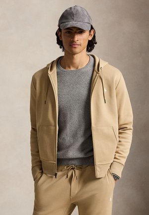 Jeune homme portant un sweat à capuche beige zippé, un pantalon de survêtement assorti, un pull gris à col rond et une casquette de baseball grise, debout devant un fond neutre.