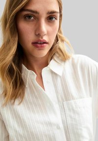 Camisa blanca de botones con rayas verticales, cuello y un bolsillo en el pecho izquierdo, hecha de una tela ligera con una textura suave.