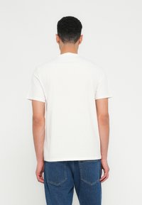 Άνδρας με κοντά σγουρά μαλλιά, φορώντας απλό λευκό μπλουζάκι κοντού μανικιού και μπλε τζιν, με την πλάτη προς την κάμερα σε λευκό φόντο.