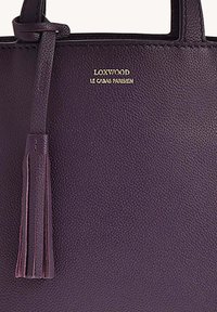 Sac fourre-tout en cuir violet avec une texture granuleuse. Présente un logo doré et une décoration en gland. Design simple et minimaliste avec des poignées courtes.