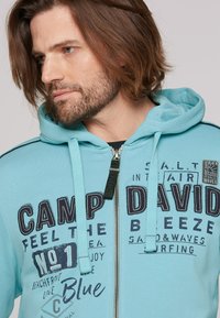 Turquoise hooded sweatshirt with zip closure, featuring navy printed text and graphics, drawstring hood, and textured fabric.