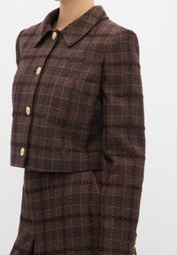 Brun ternet jakke har et kort design, en klassisk krave og guldfarvede knapper med tekstureret stof og kontrasterende ternede linjer.