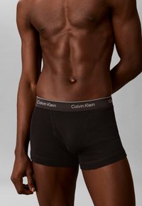 Αρσενικός κορμός που φοράει μαύρα εσώρουχα Calvin Klein με λογότυπο στη μέση, πάνω σε απλό γκρι φόντο.