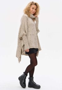 Beżowy, oversize sweter dzianinowy z wysokim kołnierzem, prążkowanymi rękawami i teksturowanym wzorem, w zestawieniu z ciemną spódnicą i czarnymi butami wojskowymi.