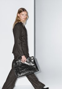 Donna in un completo scuro a righe, che cammina, tenendo una grande borsa in pelle nera strutturata con la mano destra, su uno sfondo chiaro e semplice.