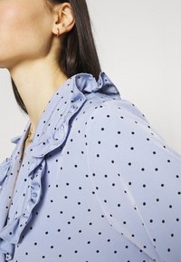Blusa azzurra con pois neri, caratterizzata da un colletto arricciato e una texture liscia. Accentuata da un delicato orecchino a cerchio in oro.
