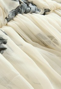 Leichtes, transparentes Gewebe mit einer cremefarbenen Basis und grauen Blumenmustern. Das Material wirkt strukturiert mit sanften, fließenden Falten.