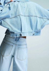 Ljusblå jeansjacka och höga jeans i midjan. Jackan är kort, med knappstängning och synliga sömmar. Jeansen har bakfickor.