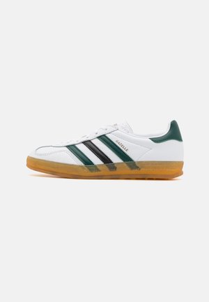 Biały sneaker Adidas Gazelle z zielonymi i czarnymi paskami, gumową podeszwą i zieloną zakładką na pięcie, pokazany z profilu na białym tle.
