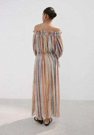 Mujer con moño vestida con un vestido largo, sin los hombros al descubierto, con rayas verticales multicolores y zapatos planos negros, de pie frente a una pared blanca.