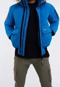 Man i en ljusblå vinterjacka med pälsluva, svart skjorta och olivgröna cargobyxor, stående med händerna i jackfickorna.