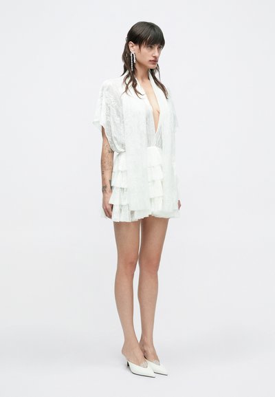 Biele vrstvené šaty s hlbokým výstrihom do tvaru V, krátkymi rukávmi a priehľadnou, textúrovanou látkou. Kombinované s bielymi topánkami s špicatou špičkou.