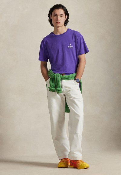 T-shirt en coton violet avec logo, pantalon blanc, pull vert noué autour de la taille, et baskets orange avec accents jaunes. Design simple et décontracté.