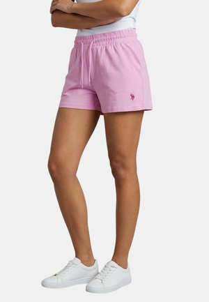 Frau trägt hellrosa Shorts mit Kordelzug und weiße Turnschuhe, steht mit verschränkten Armen vor einem einfarbigen Hintergrund.