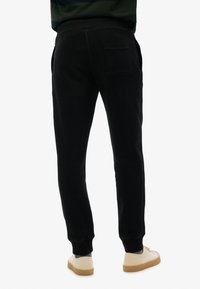 Zwarte joggingbroek met een zachte textuur, elastische tailleband, taps toelopende benen en een achterzak. Gekoeld met lichtgekleurde schoenen.