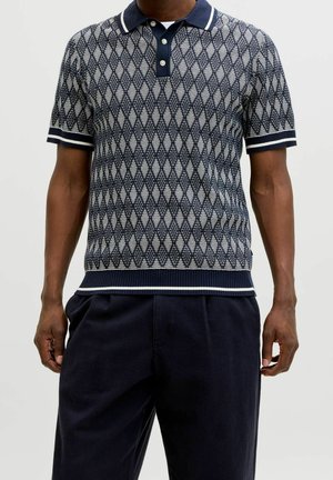 Homme portant un polo en maille à manches courtes bleu marine et blanc à motif losanges et un pantalon bleu marine foncé sur fond uni.