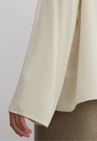 Helle beige Bluse mit langen Ärmeln, aus glattem Stoff gefertigt, mit lockerer Passform, weiten Ärmeln und klaren Saumdetails.