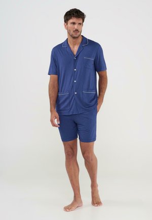 Hombre de pie descalzo vistiendo una camisa azul de manga corta con botones y pantalones cortos a juego, con detalles de ribete blanco y bolsillos.