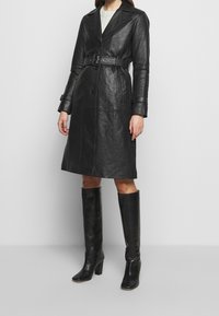 Black leather trench coat with a belted waist, wide lapels, and button closure, paired with knee-high black heeled boots.